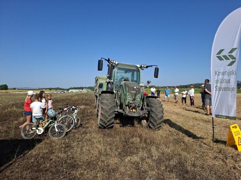 Sodoben traktor na njivi, poleg pa so otroci s kolesi.